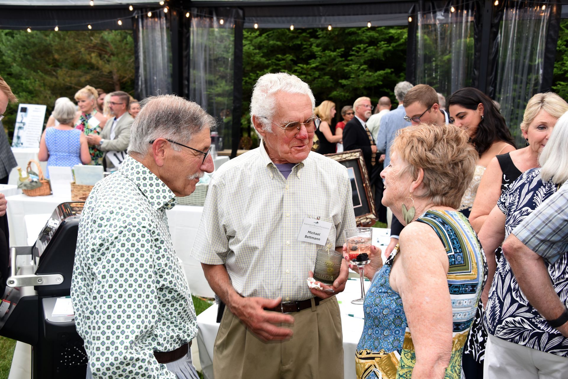 Group of older adults at an outdoor event, conversing near a table. People are dressed in summer attire.