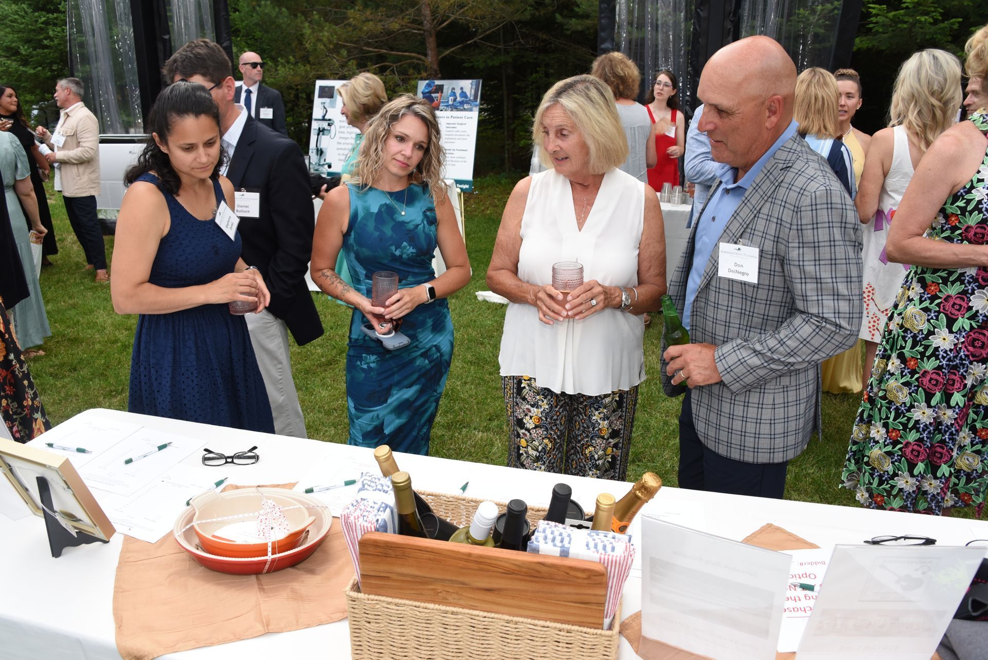 Group of people gathered at an outdoor event with a table displaying items. Women and men are holding drinks.