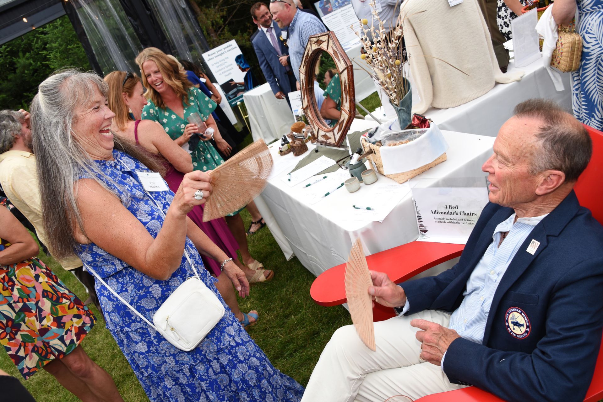 Woman in blue dress laughs, holding a fan, gesturing towards a man in a blazer seated in a red chair at an outdoor event.