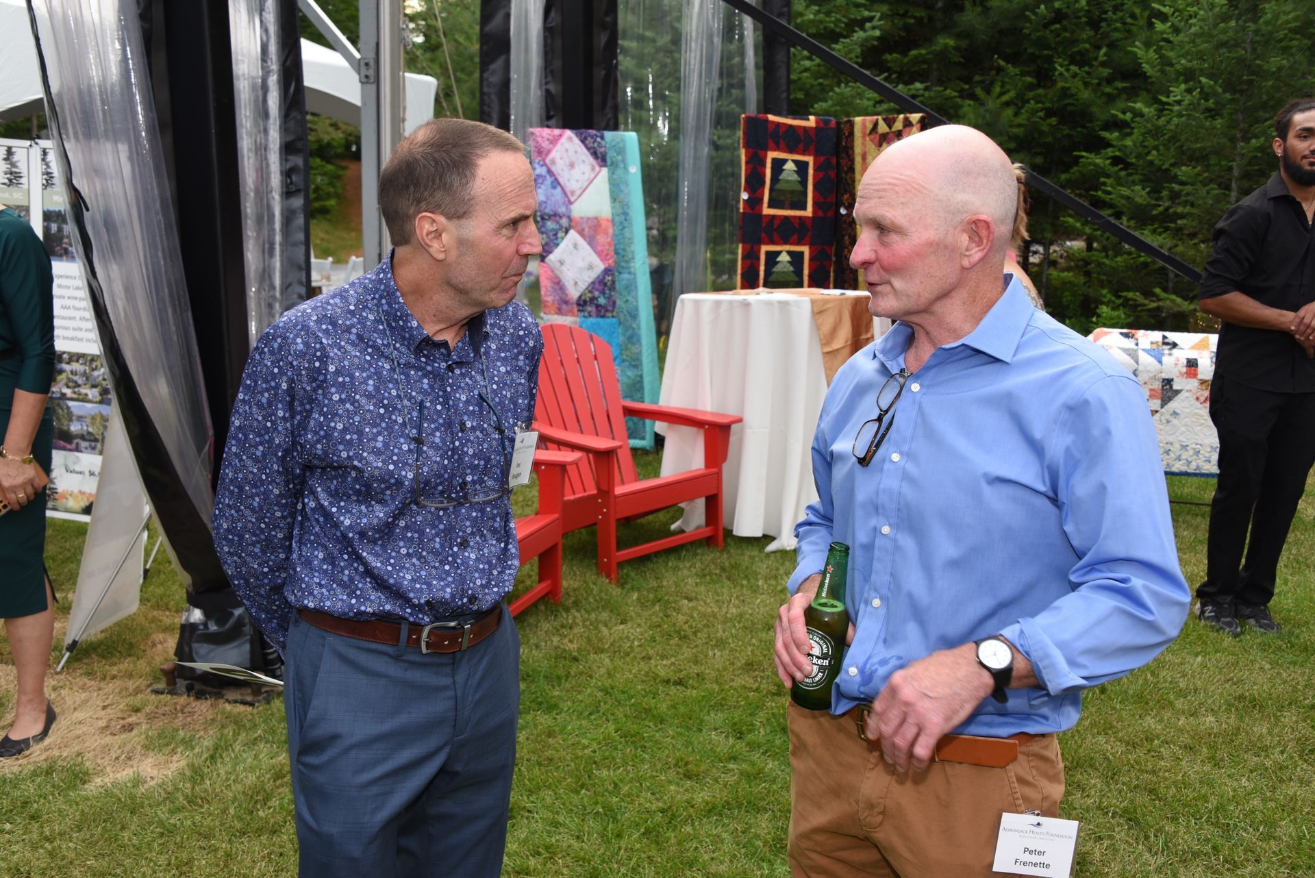Two men in a grassy outdoor setting converse. One in a blue floral shirt and the other in a light blue shirt and khaki pants.