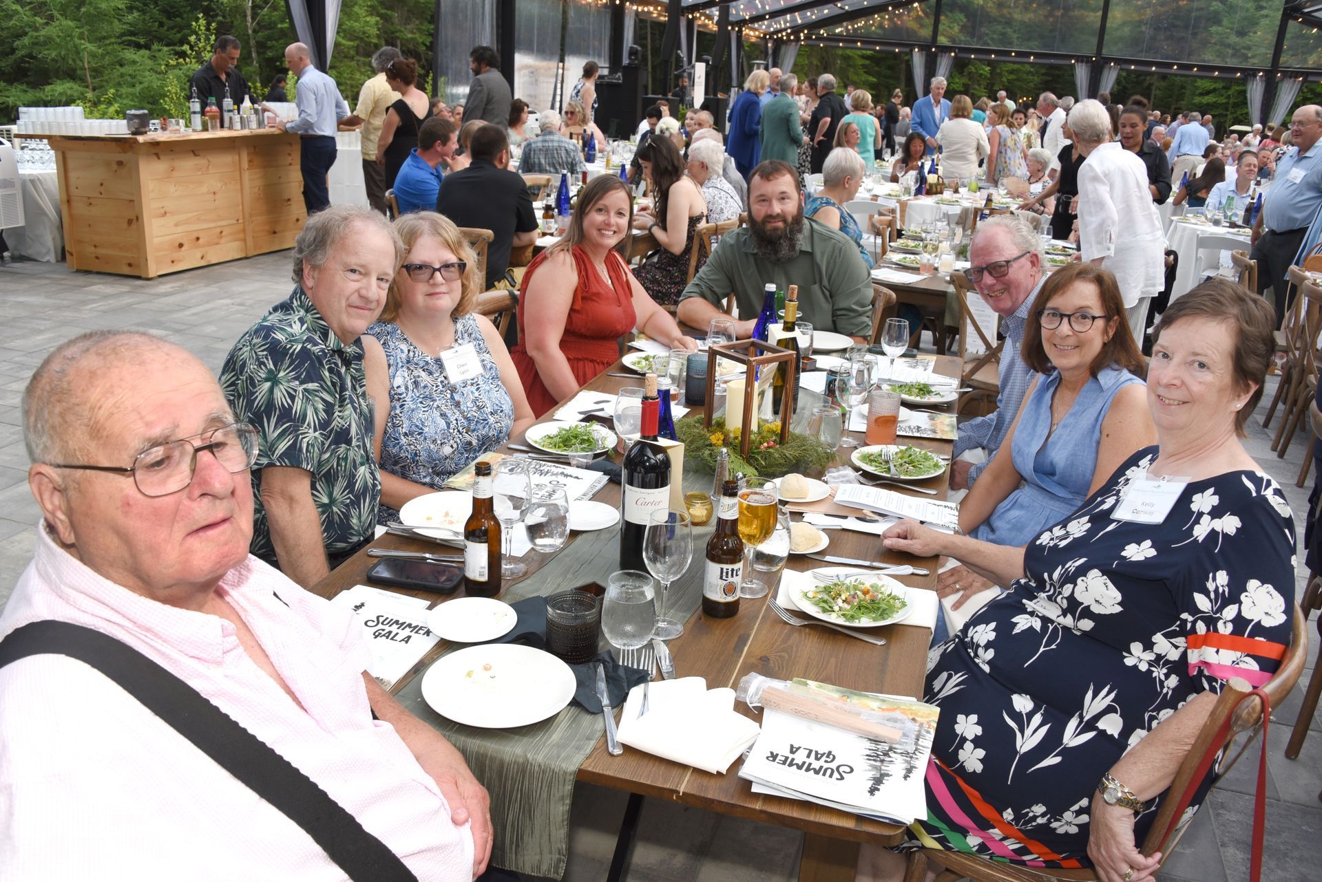 People seated at a long outdoor table, enjoying a meal at an event. Guests are smiling and conversing.