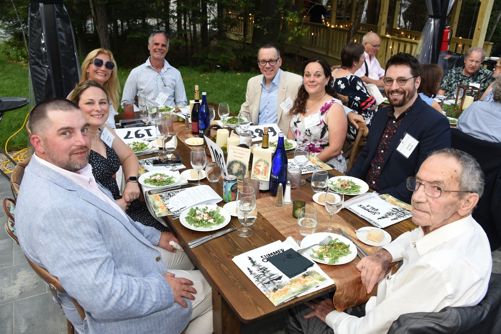 People seated at a long wooden table outdoors, enjoying a meal. Plates of food and bottles of wine are visible