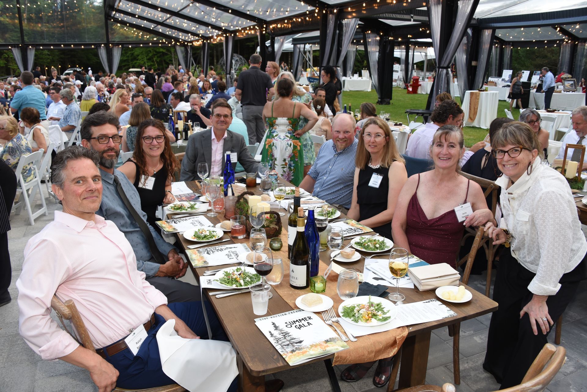 Group of people seated around a long table at an outdoor event, enjoying a meal under a transparent tent.