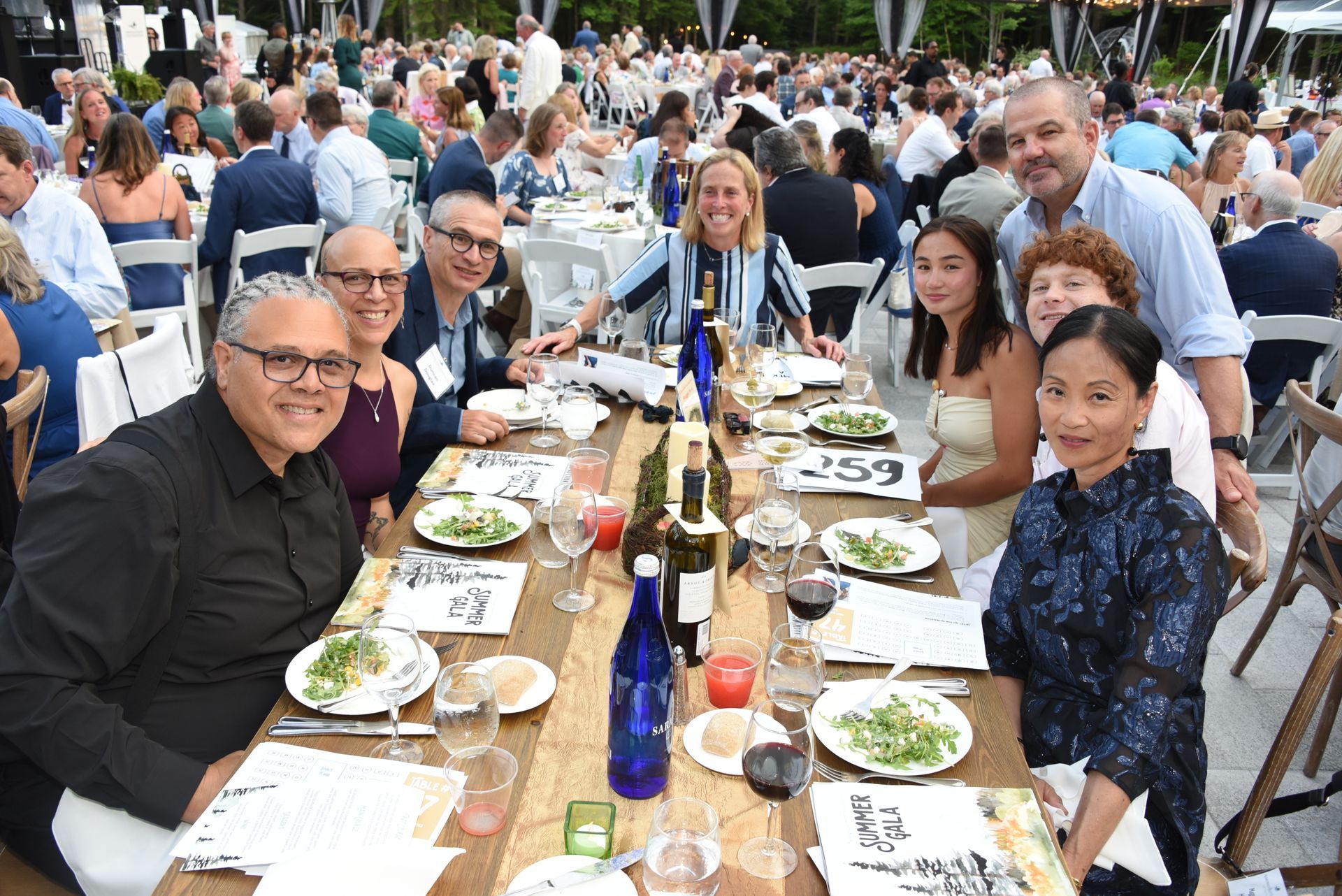 Group of people seated at a long table outdoors, smiling and eating. Plates of food, wine bottles.
