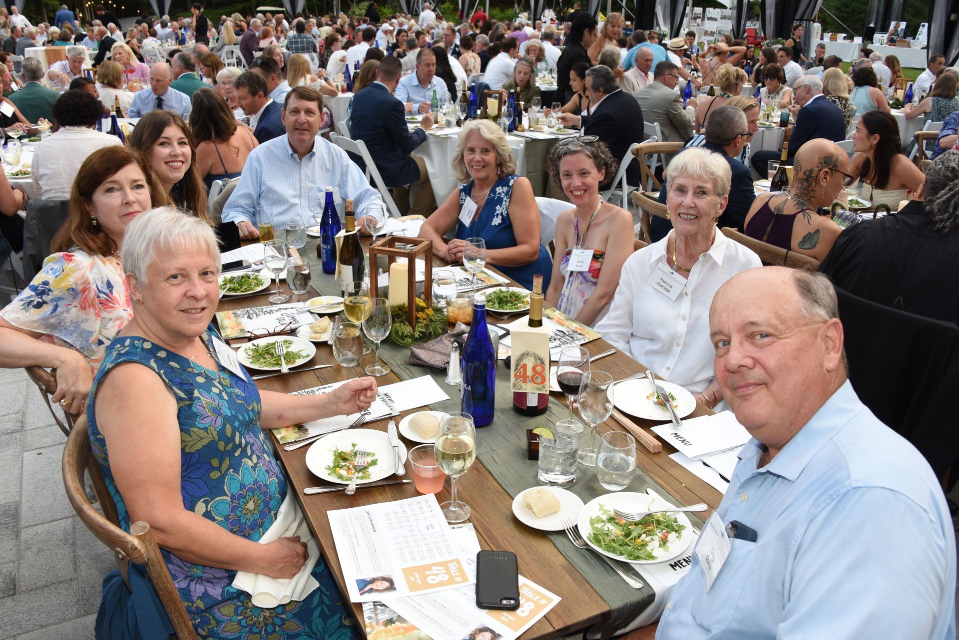 People seated at a large outdoor dinner party; tables with food and wine. 