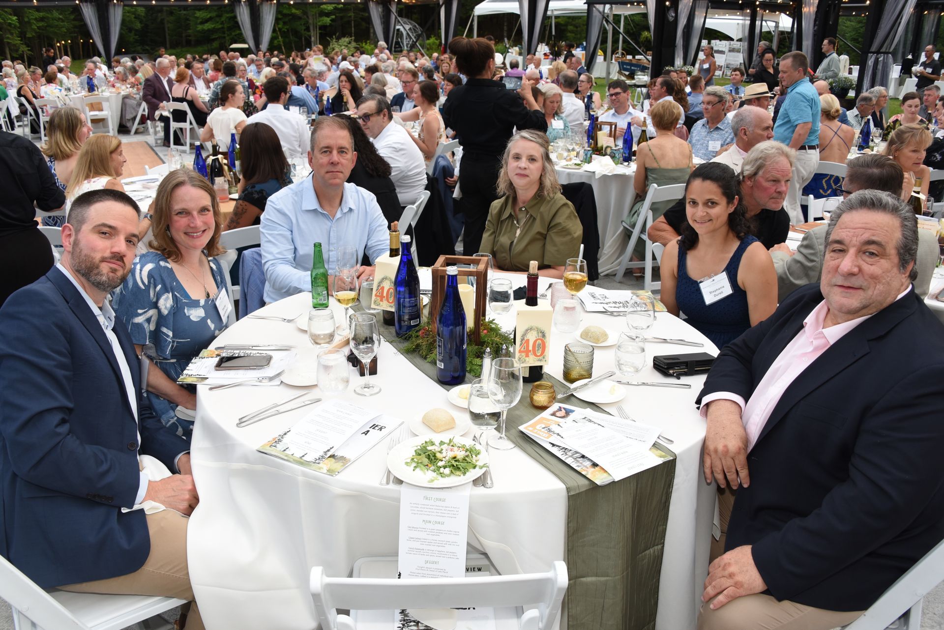 A group of people seated at a round table at an outdoor event, smiling at the camera. They are dining and socialising.