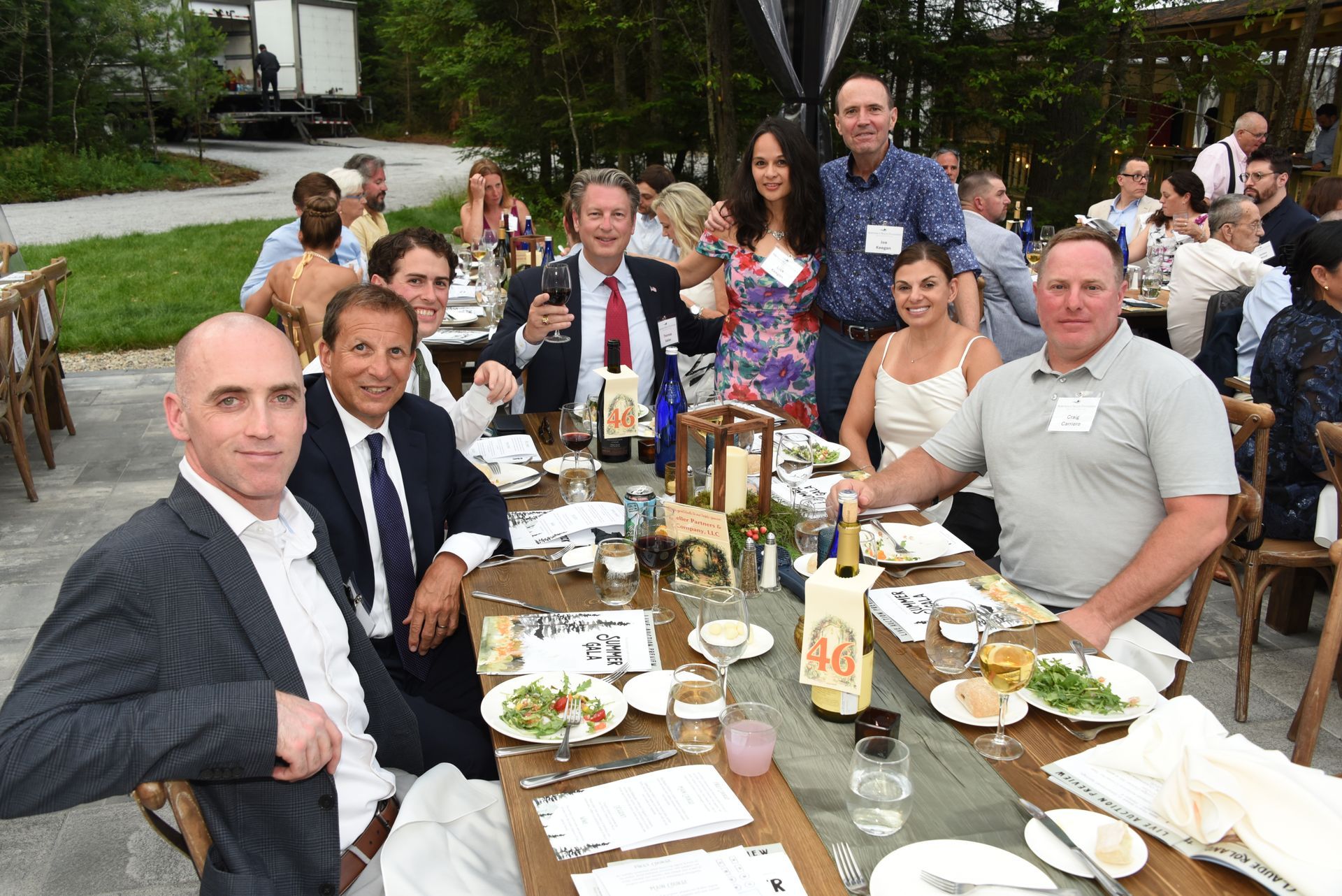 A group of people seated around a long table outdoors, smiling and enjoying a meal.