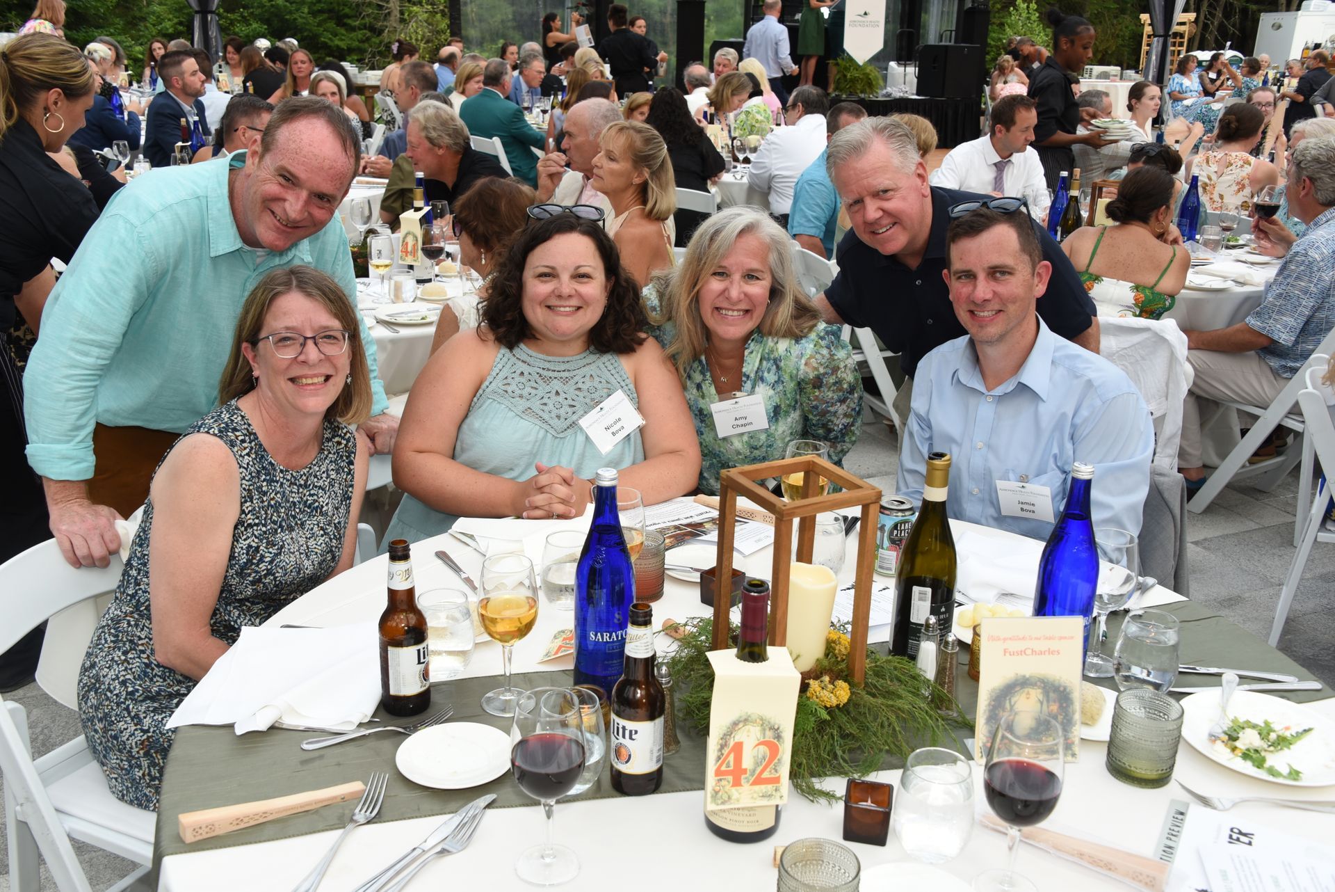Group of smiling people seated at a table outdoors, likely at a social gathering.