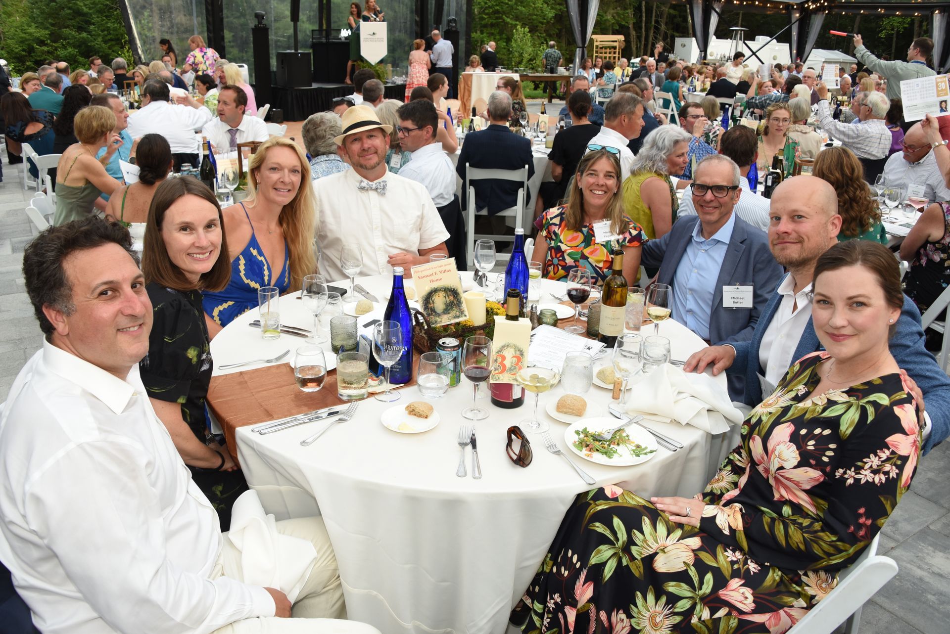 Group of people seated around a white table at an outdoor event, smiling. Some are looking at the camera.