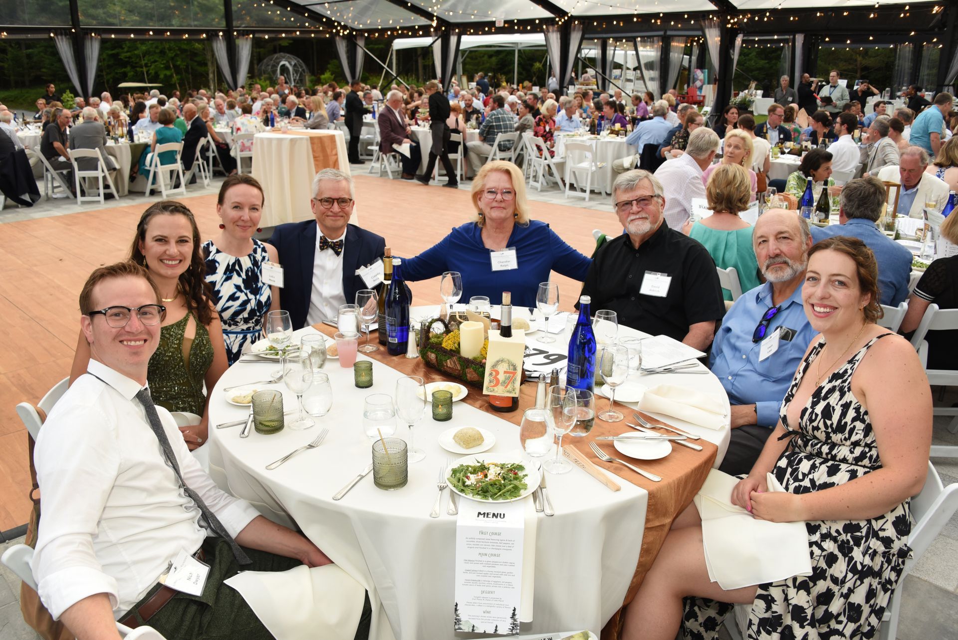 Group of people seated at a round table at an outdoor event, smiling at the camera.