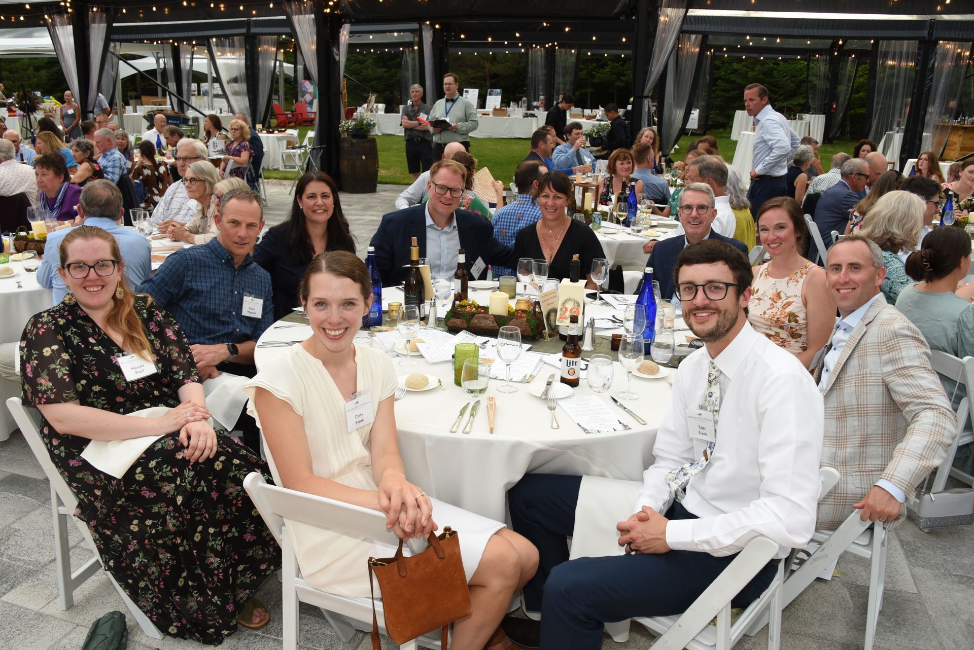 A group of people at a formal outdoor event, sitting around a table. Attendees are smiling and talking.