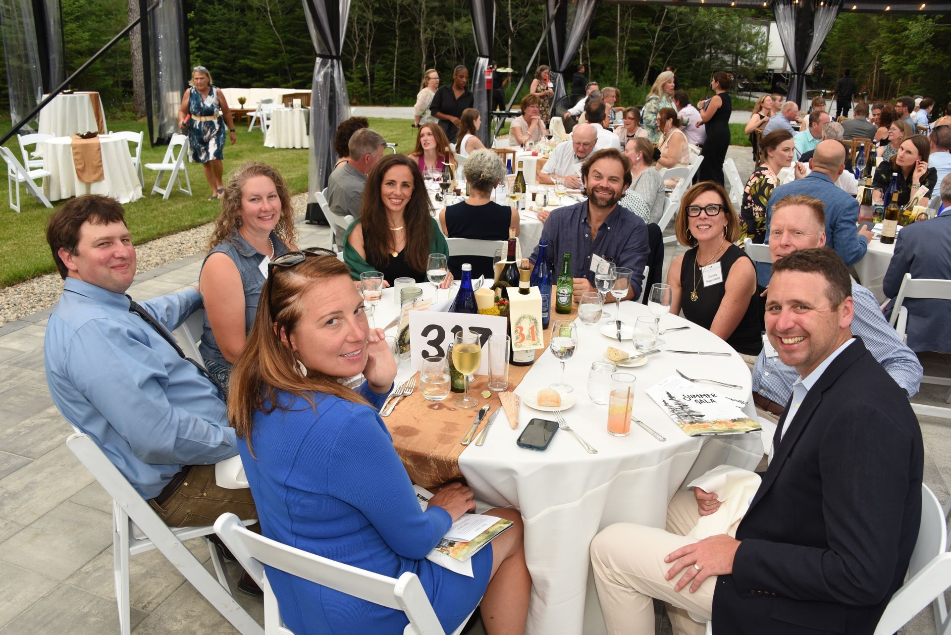 Group of people seated around a table at an outdoor event.  They're smiling, in a well-lit space with a tent.