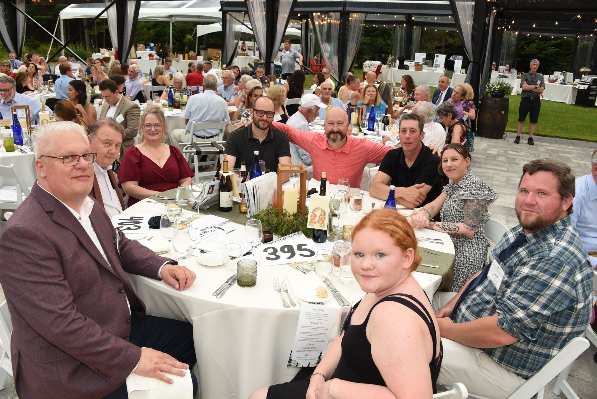 A large group of people seated at tables at an outdoor event, many smiling and facing the camera.