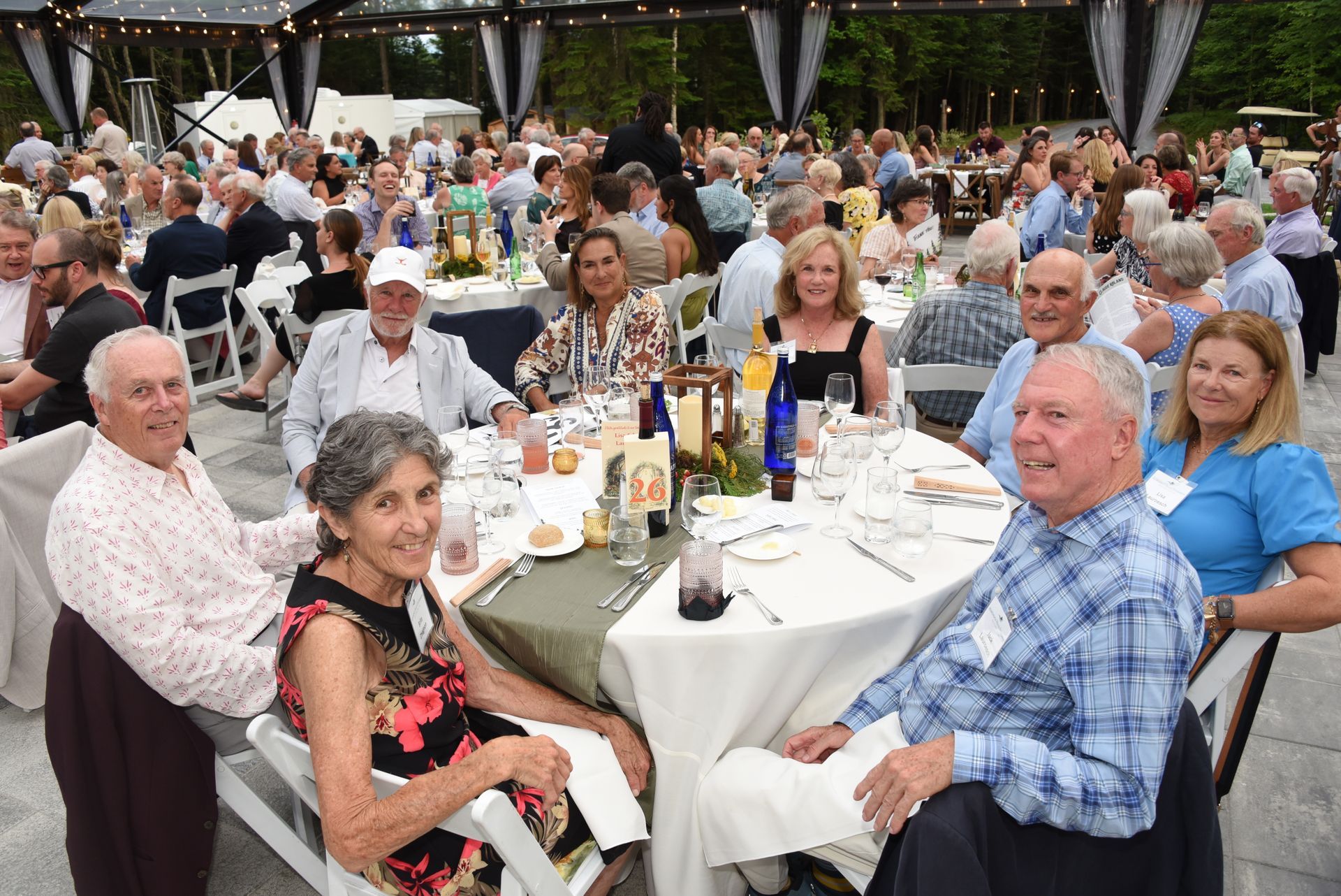 People seated at tables at an outdoor event, likely a dinner, with many guests in the background. Some smiling at the camera.