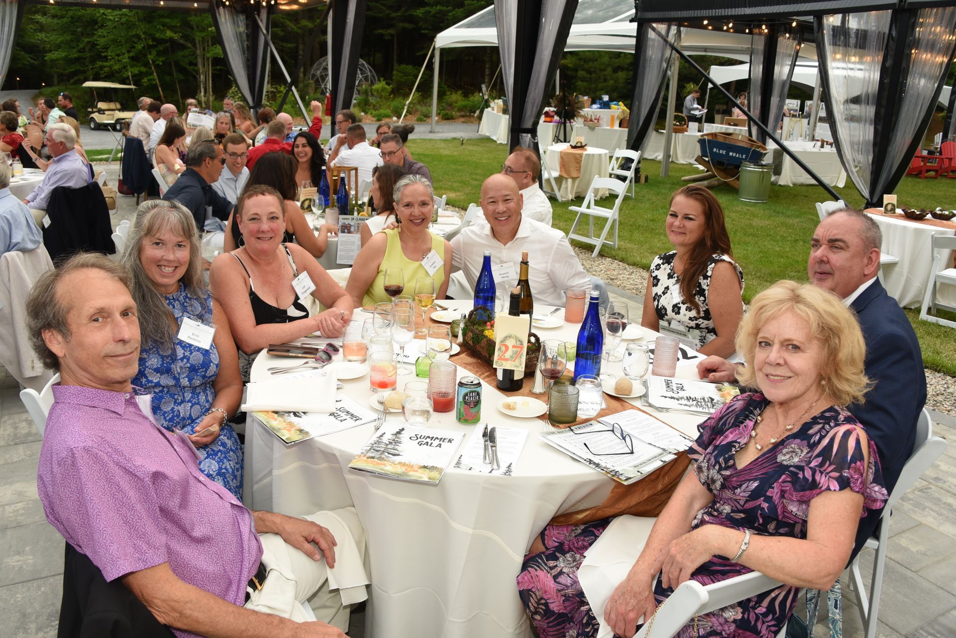 A group of people seated at a round table at an outdoor event, smiling and socializing.