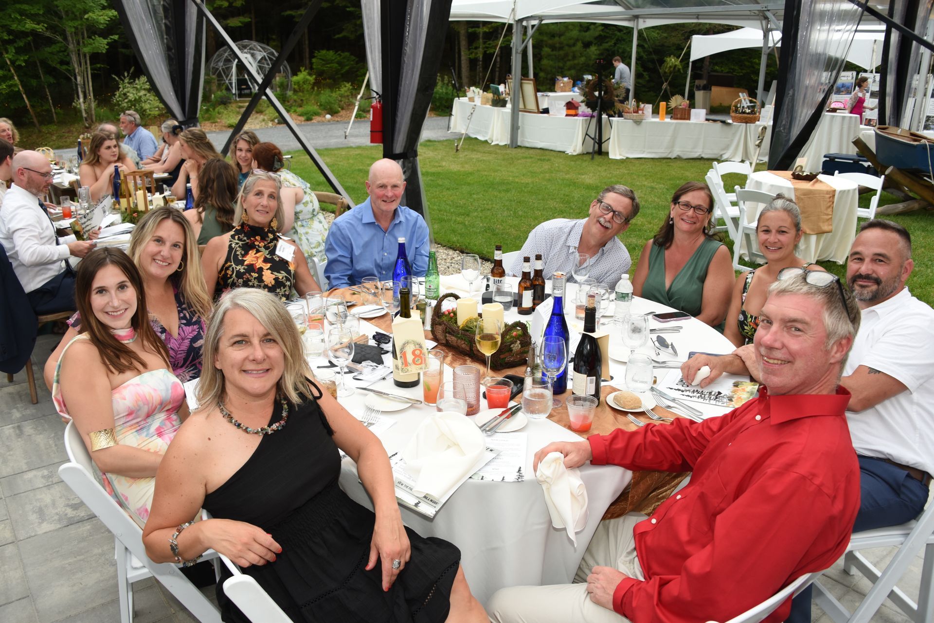 Group of people at a table set for an outdoor event. People smile and pose, in front of a backdrop of tents and greenery.