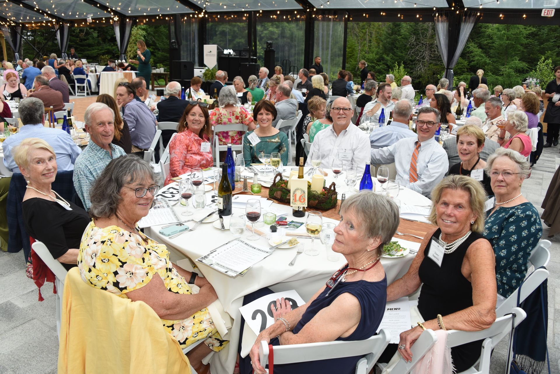 A well-attended outdoor event with people seated at tables. Guests are dressed up and enjoying a meal under a clear tent.