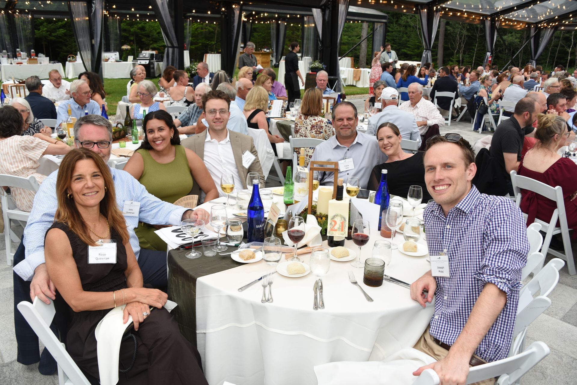 A group of people seated at a round table at an outdoor event, smiling and looking at the camera.