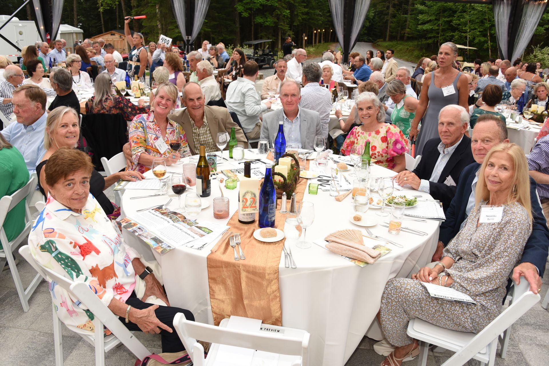 Group of people seated at a round table outdoors, enjoying a meal. White tablecloth, gold runner, and flowers.