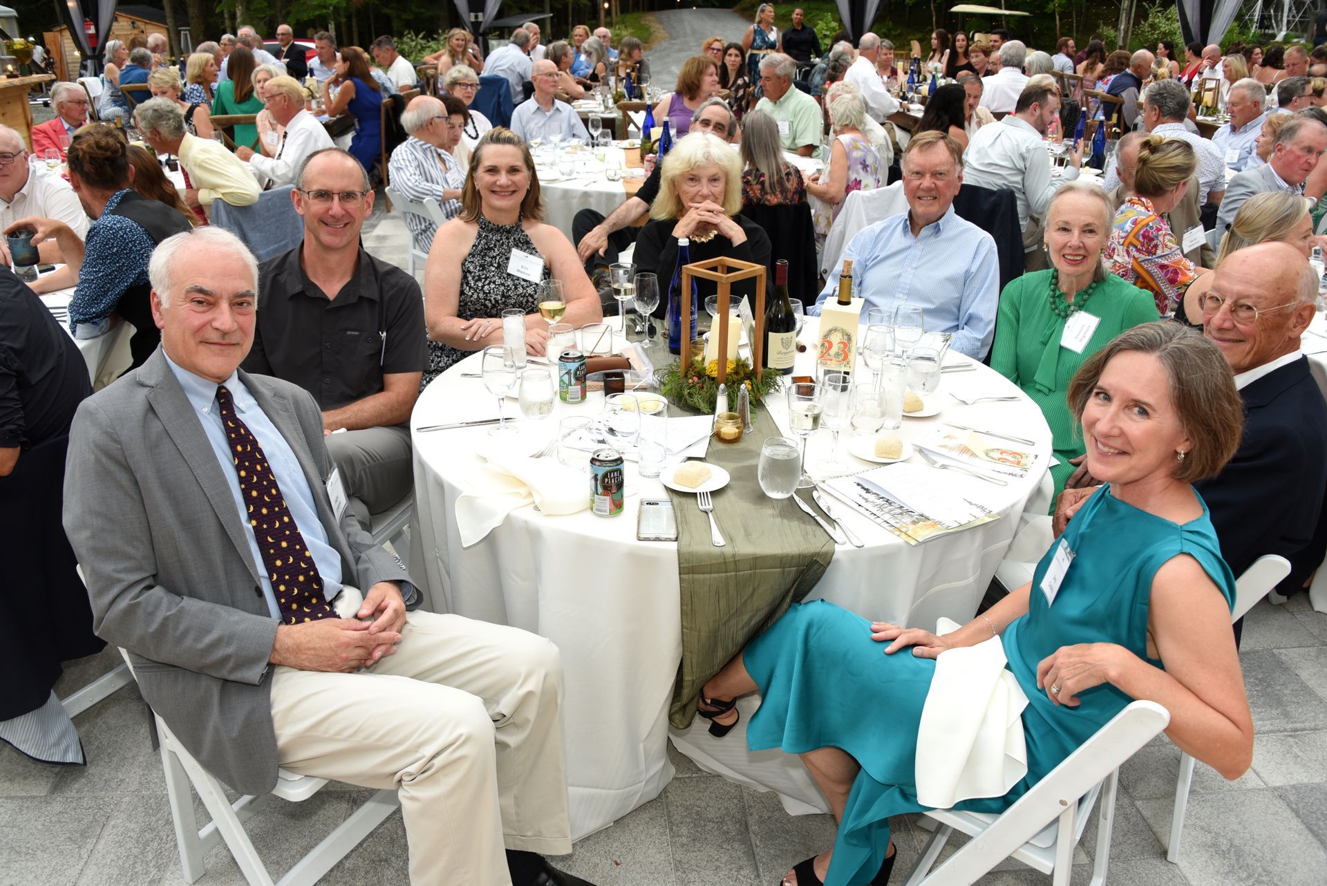 A group of people seated at a round table outdoors. Some are smiling and looking towards the camera.