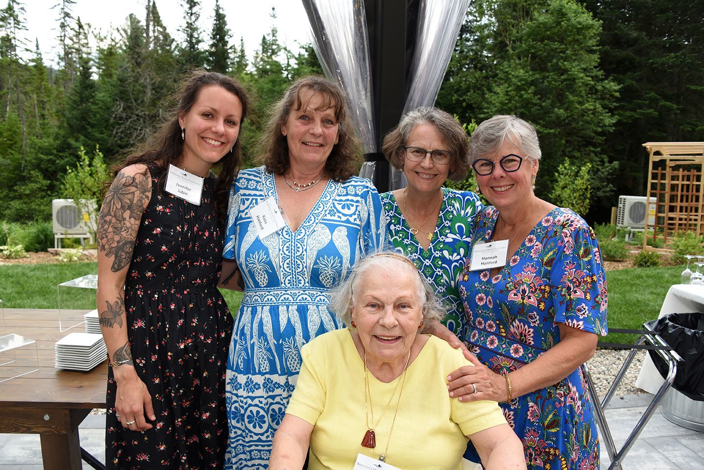 Five women, smiling, pose outdoors. An older woman is seated, surrounded by four standing women, all smiling at the camera.