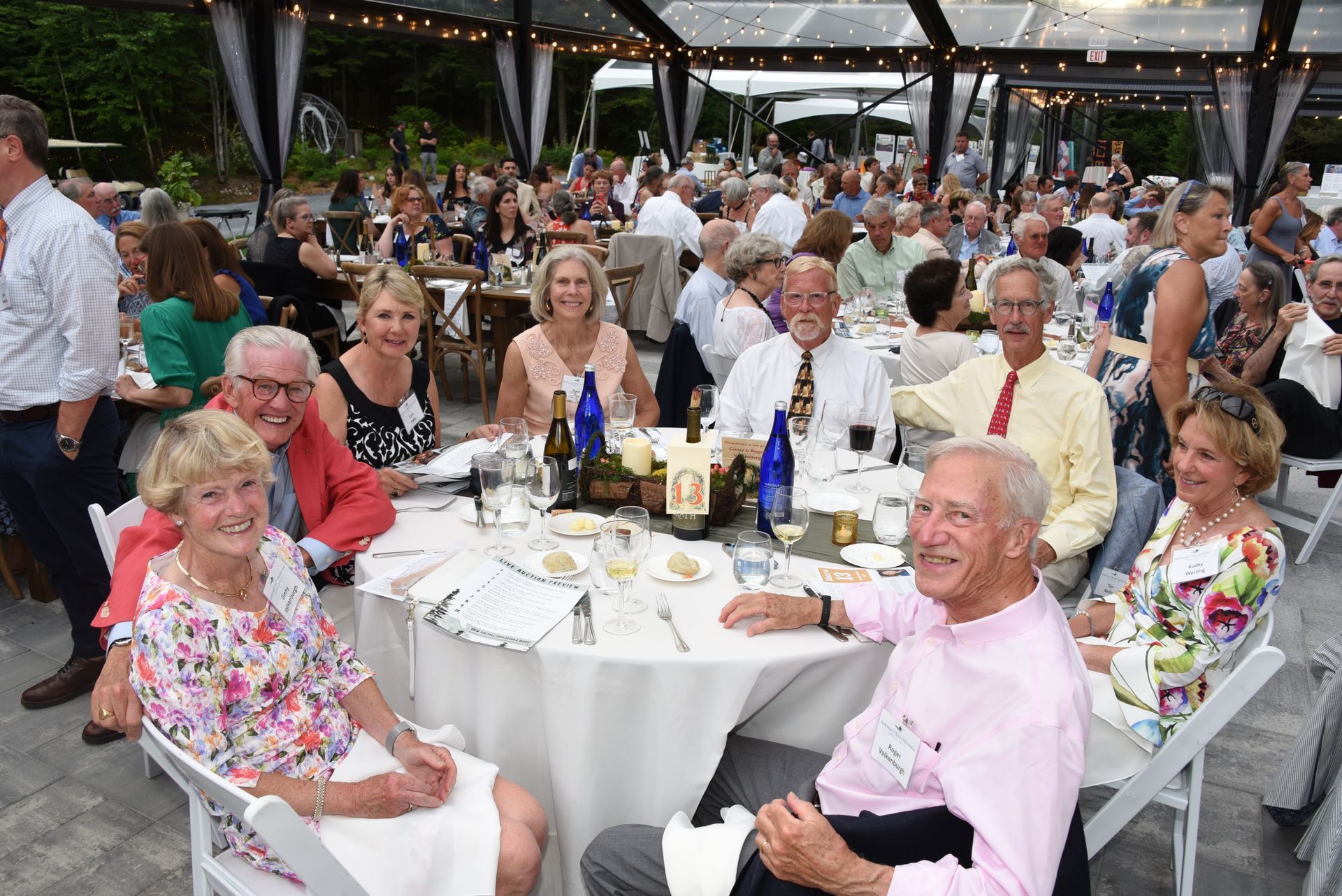 A group of people sit at a round table at an outdoor event. Many are smiling, and the setting appears to be a dinner party.