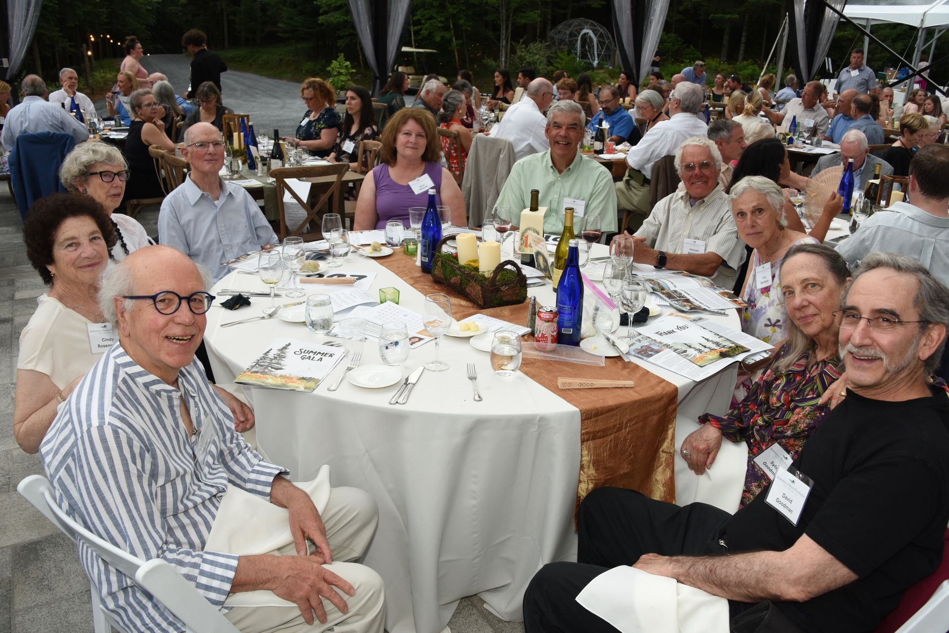 People seated at a round table outdoors at an event, with a central centerpiece. Several people are smiling.