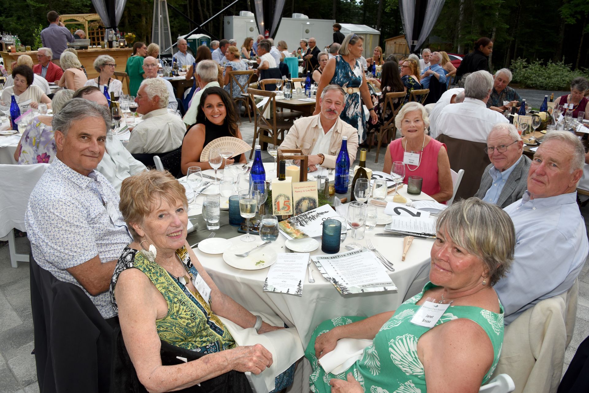 People seated around a table at an outdoor event, eating and socializing. Many are smiling and looking at the camera.
