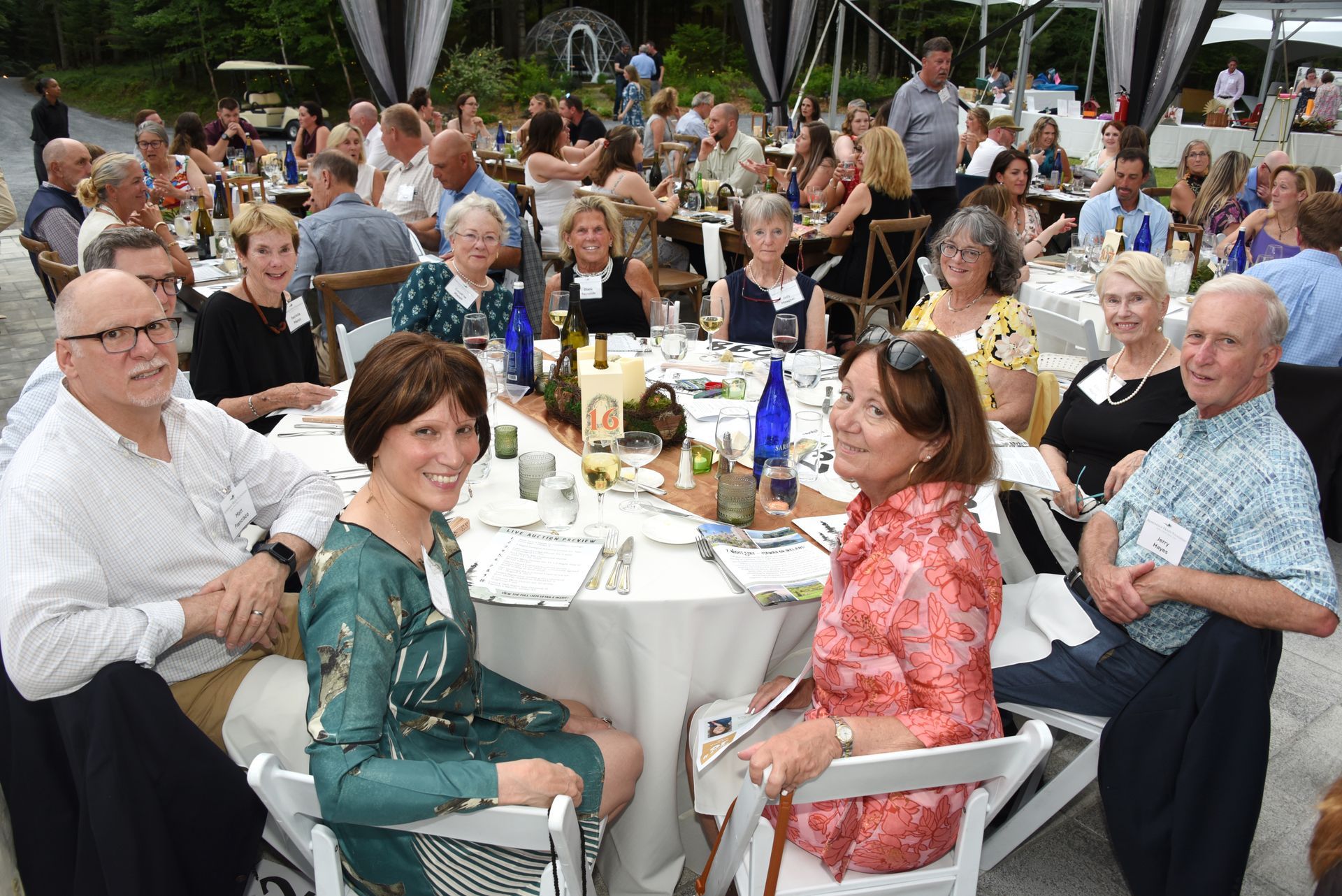 People seated at round tables outdoors, dining at an event. Tables set with food and decorations, guests smiling.