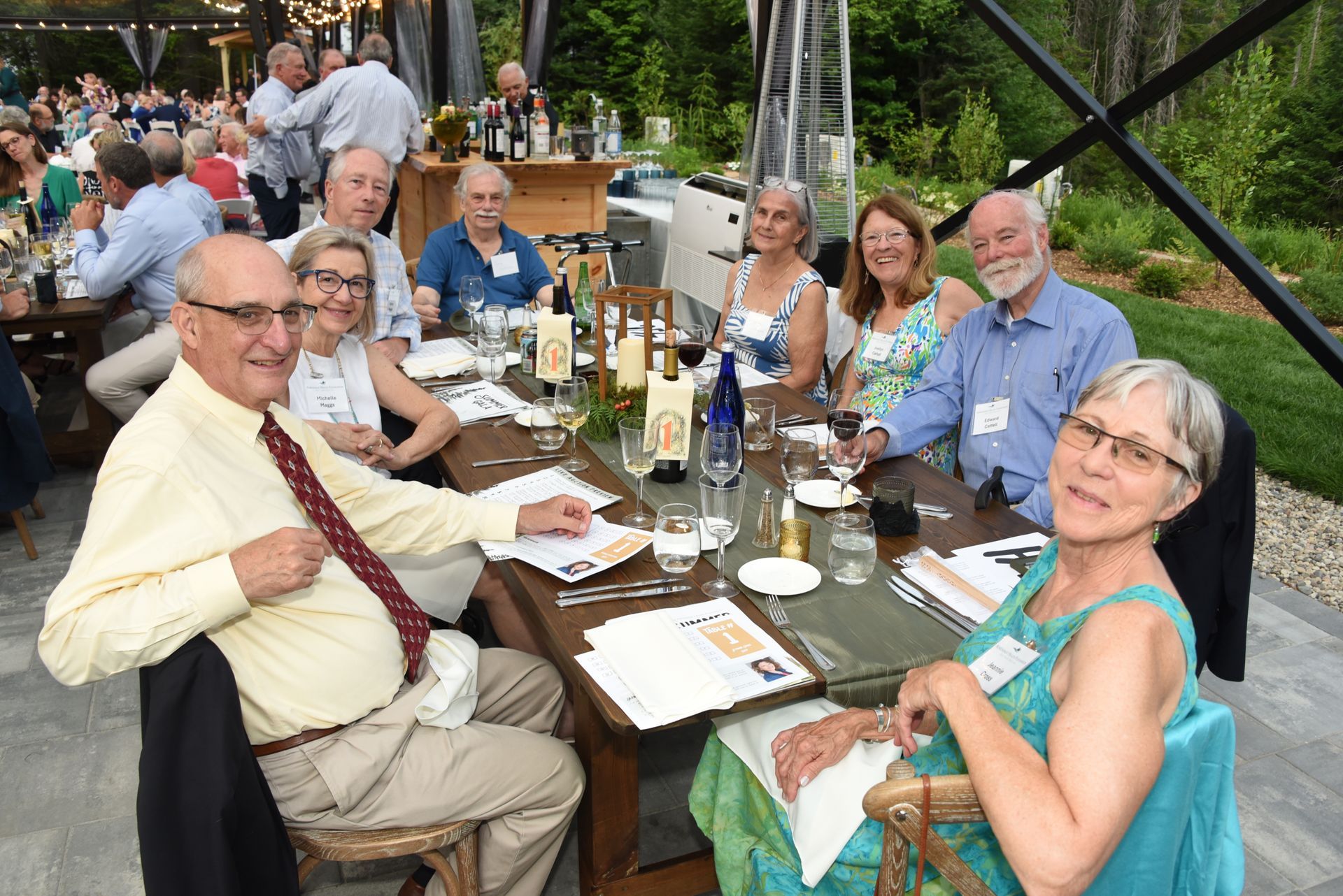 Group of people seated around a long table at an outdoor dinner, some smiling at the camera.