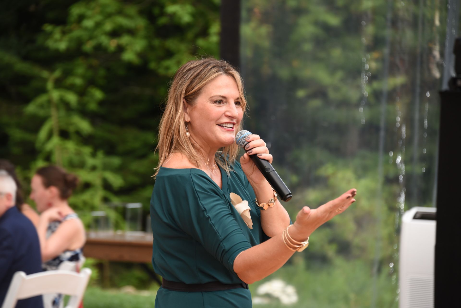 Woman in teal dress speaking into a microphone, gesturing with her right hand. Outdoors, with trees in the background.