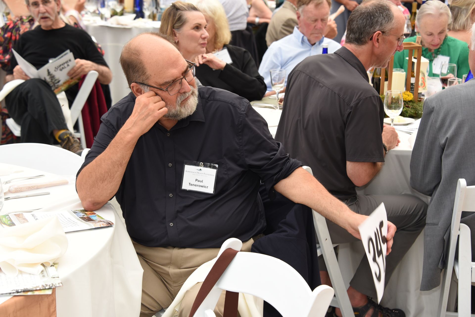 Man with a beard and glasses at a table, leaning on his hand, attending an event. He has a name tag.