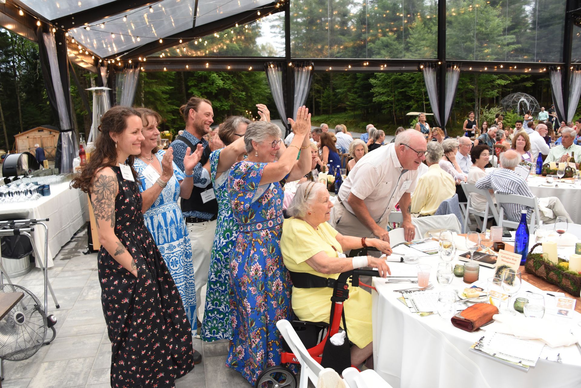 Group of people clapping at a table in a clear tent. An older woman in a wheelchair is smiling.