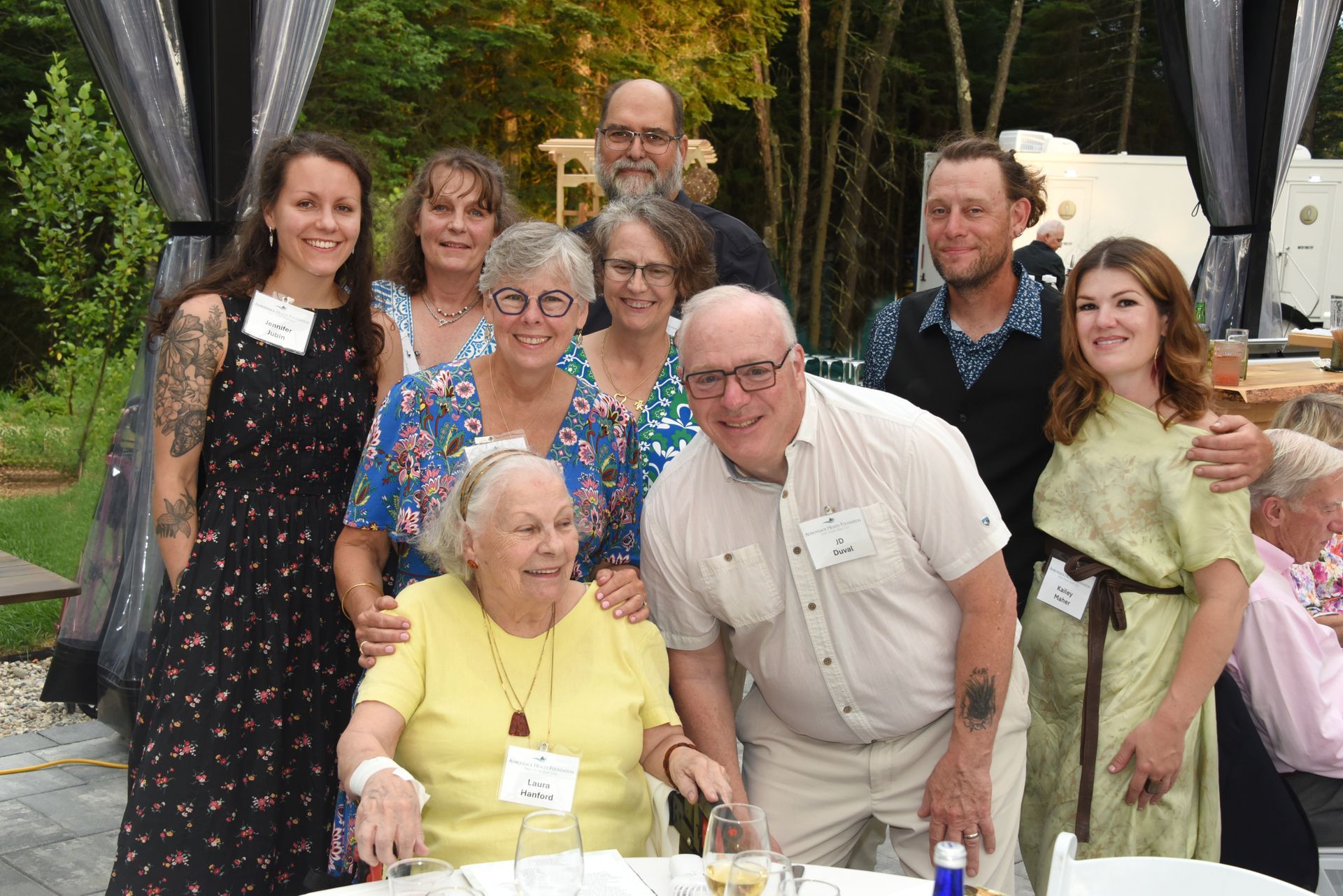 Group of people posing for a photo outdoors; diverse ages, smiling, some wearing name tags. Woman in yellow seated front.