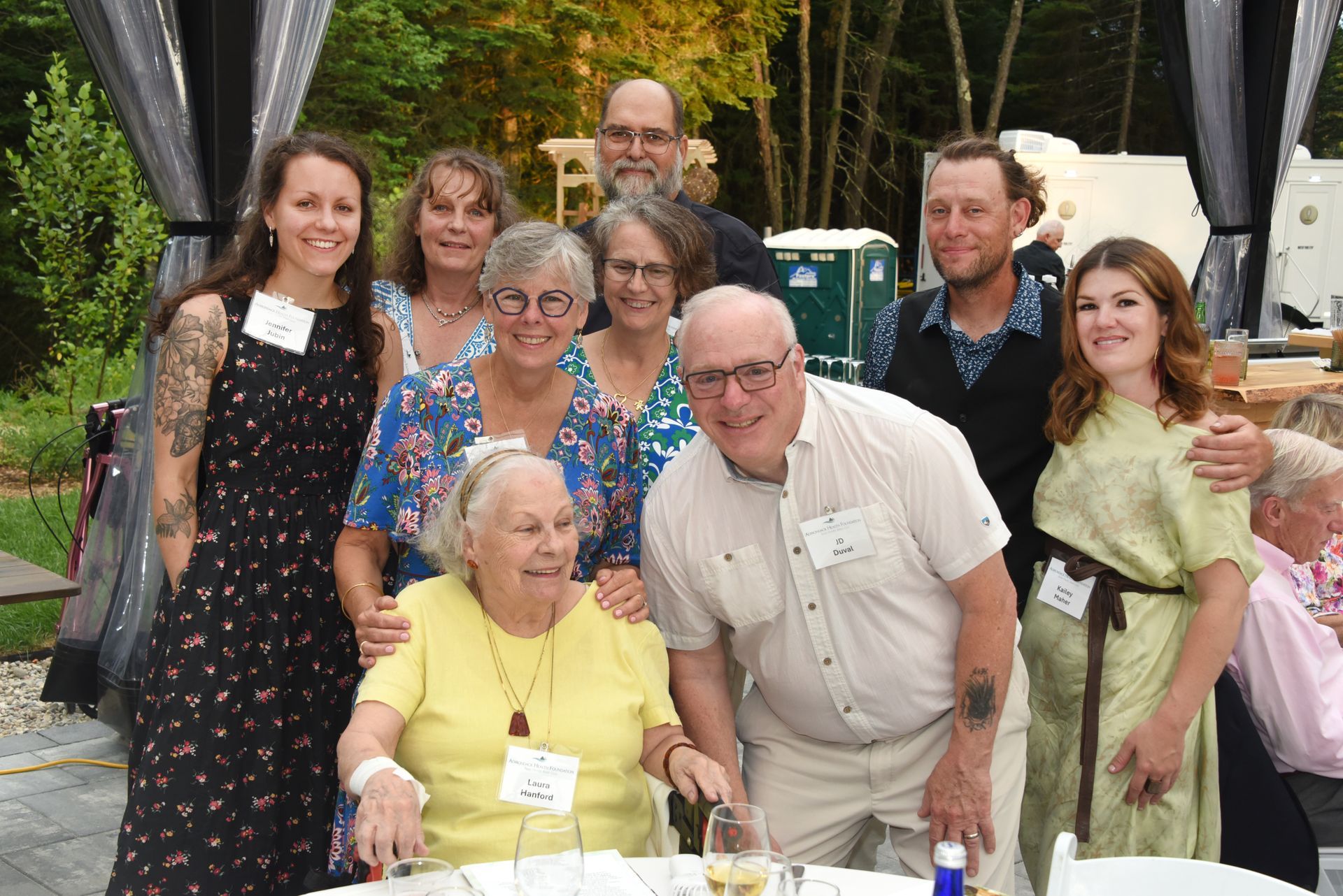 Group portrait of ten people smiling, gathered outdoors. People are of various ages and wear summer clothing.
