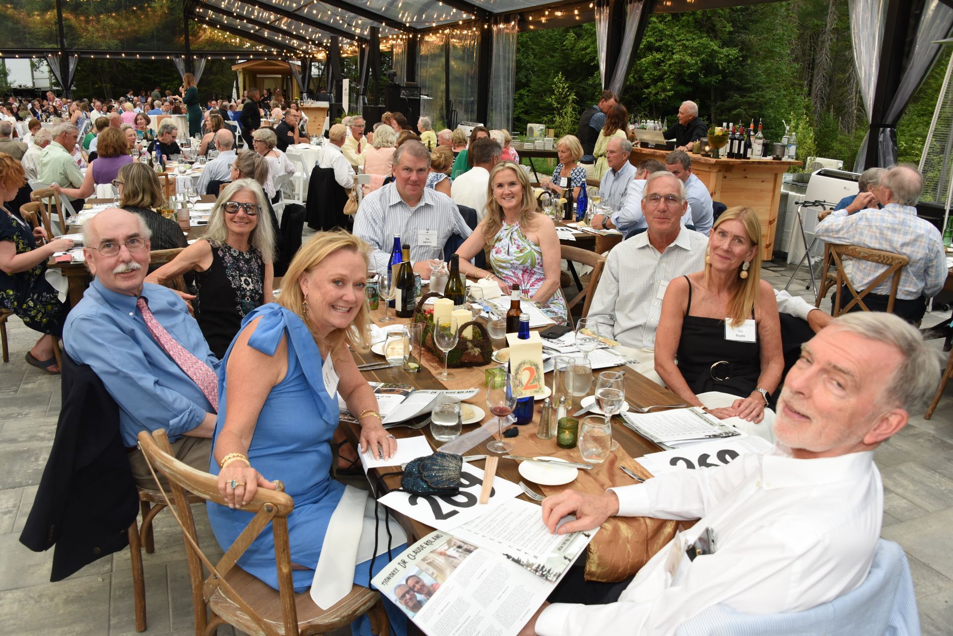 People seated at a long table at an outdoor event under a transparent roof.