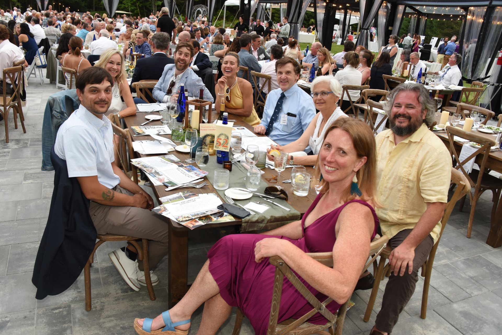 People seated at a long table outdoors, smiling and interacting. The table is set for a meal.