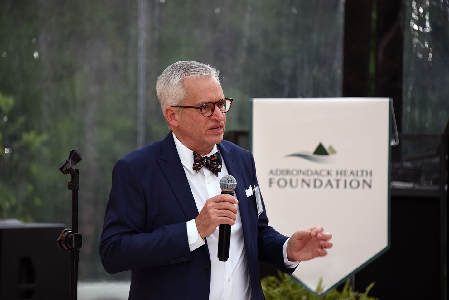 A man with glasses speaks into a microphone at an event. He stands in front of an 