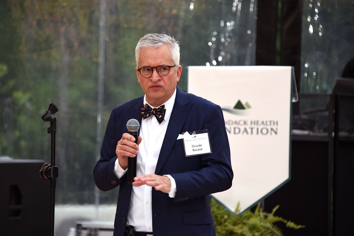 Man in a suit speaks into a microphone at an outdoor event, with a banner behind him.