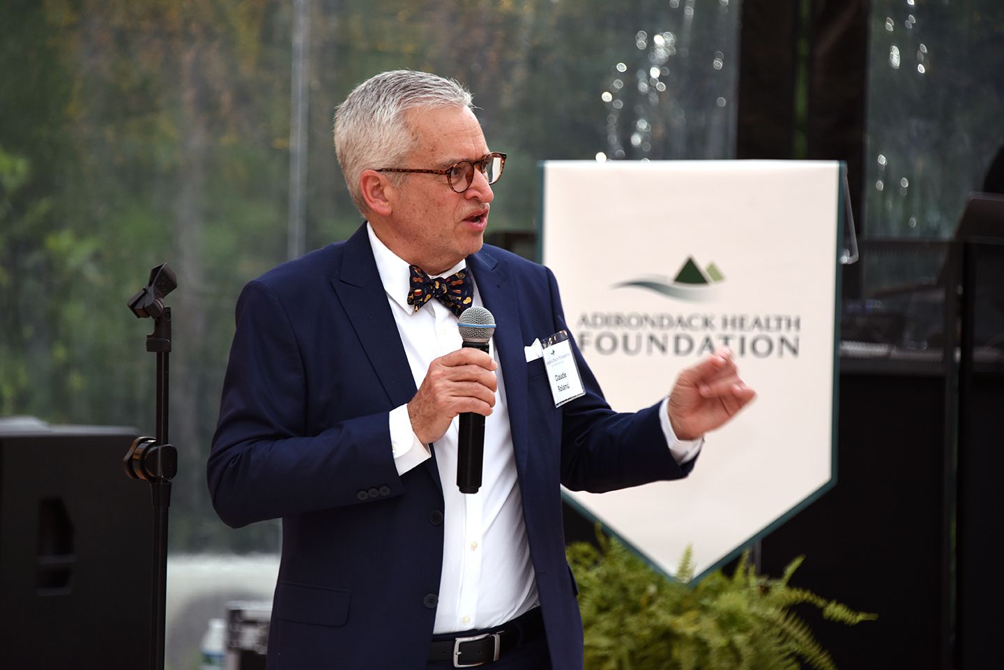 Man in a suit speaking into a microphone at an outdoor event; Adirondack Health Foundation banner visible in the background.