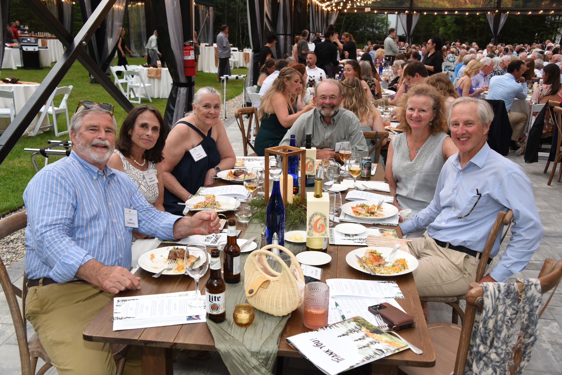Guests seated at a long table at an outdoor event; they are eating, smiling, and drinking.