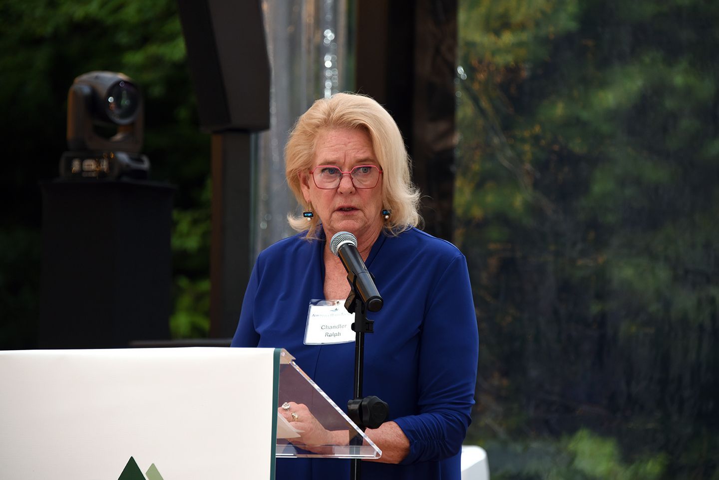 Woman with blonde hair speaking at a podium, wearing a blue blazer. Outdoors with a forest backdrop and a camera.