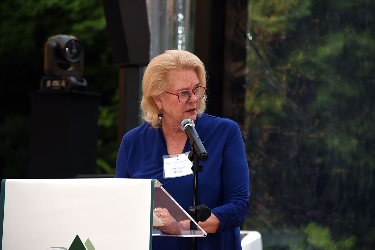 Woman with blonde hair speaks at a podium outdoors, wearing a blue top. Behind her is a backdrop of trees.