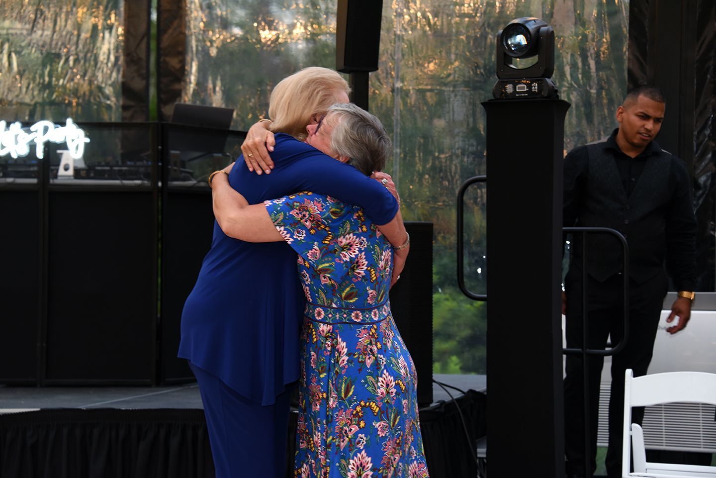 Two women embrace on a stage, one in blue, the other in a floral dress. A man in a black shirt watches nearby.
