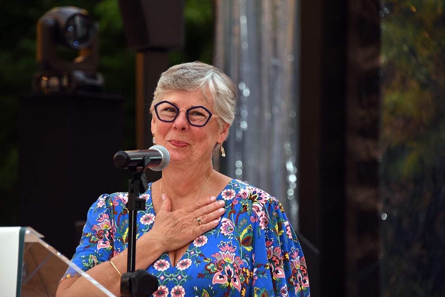 Woman with gray hair and glasses speaking into a microphone, hand over her heart, at an outdoor event.