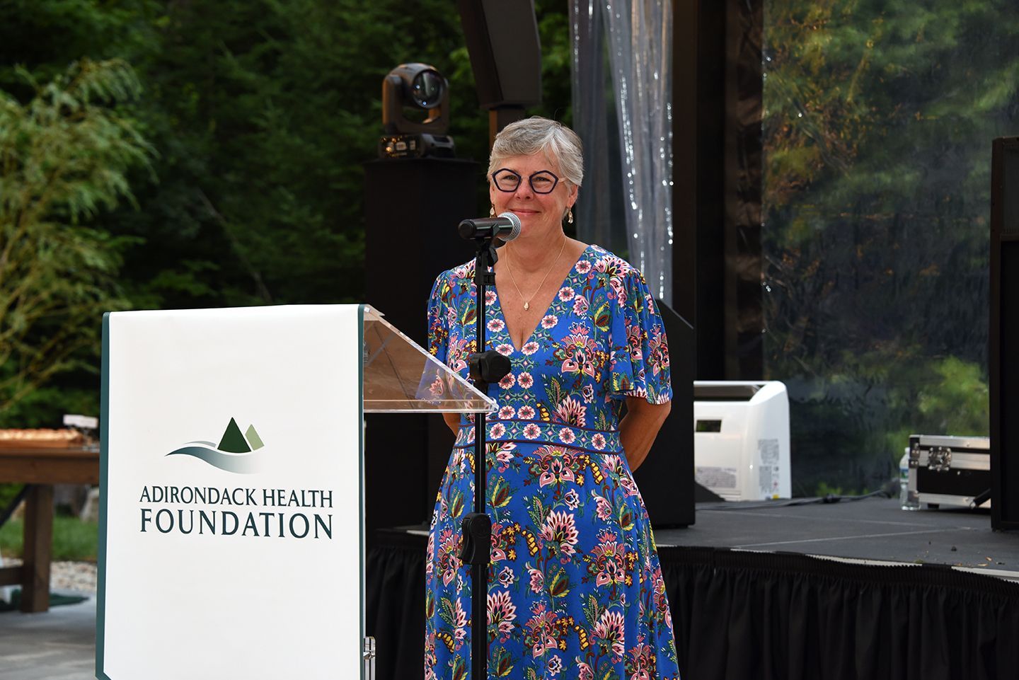 Woman in blue floral dress speaking at a podium for the Applegate Health Foundation, outdoor setting.