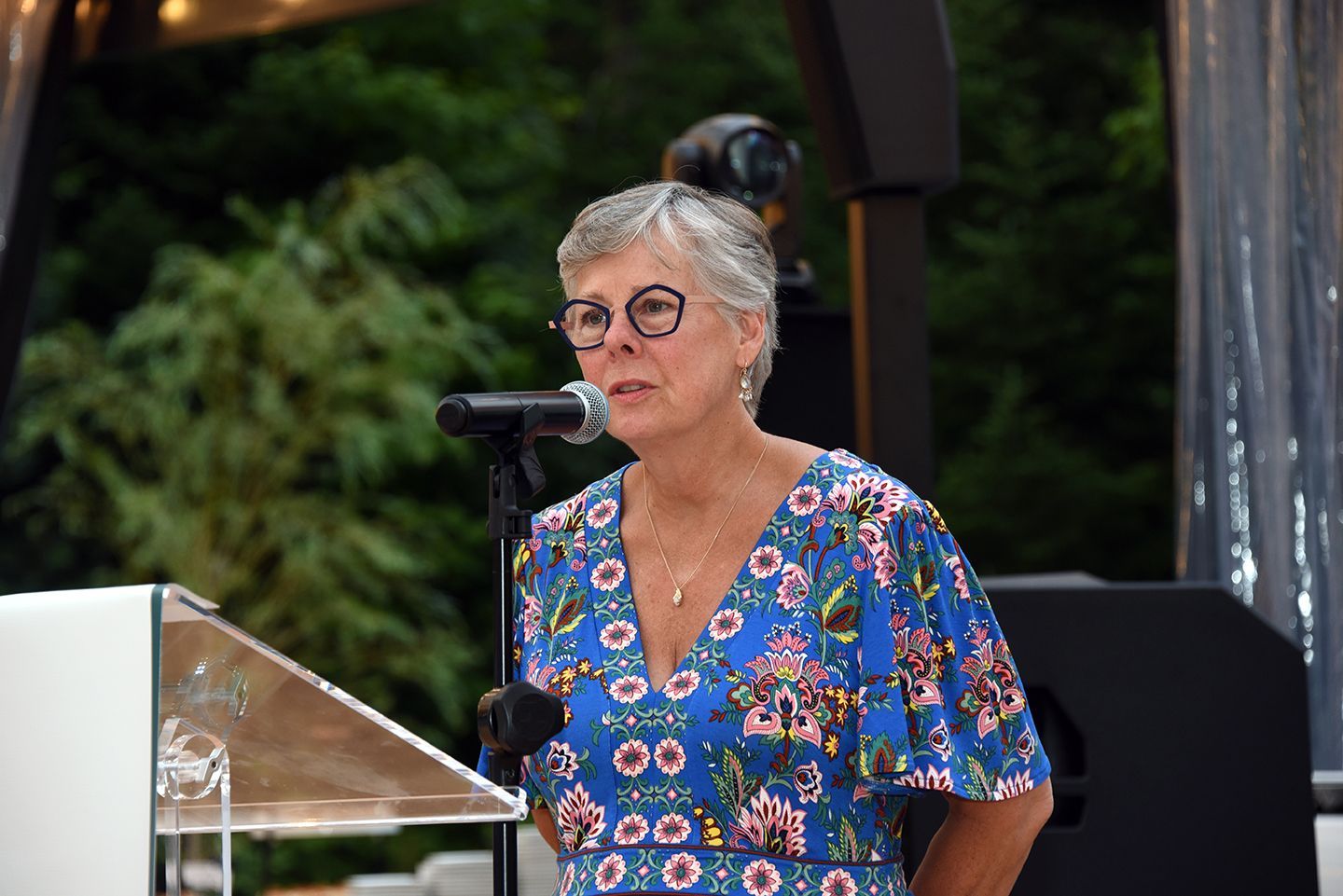 Woman speaking at a podium outdoors. She wears glasses and a blue floral dress, and is holding a microphone.