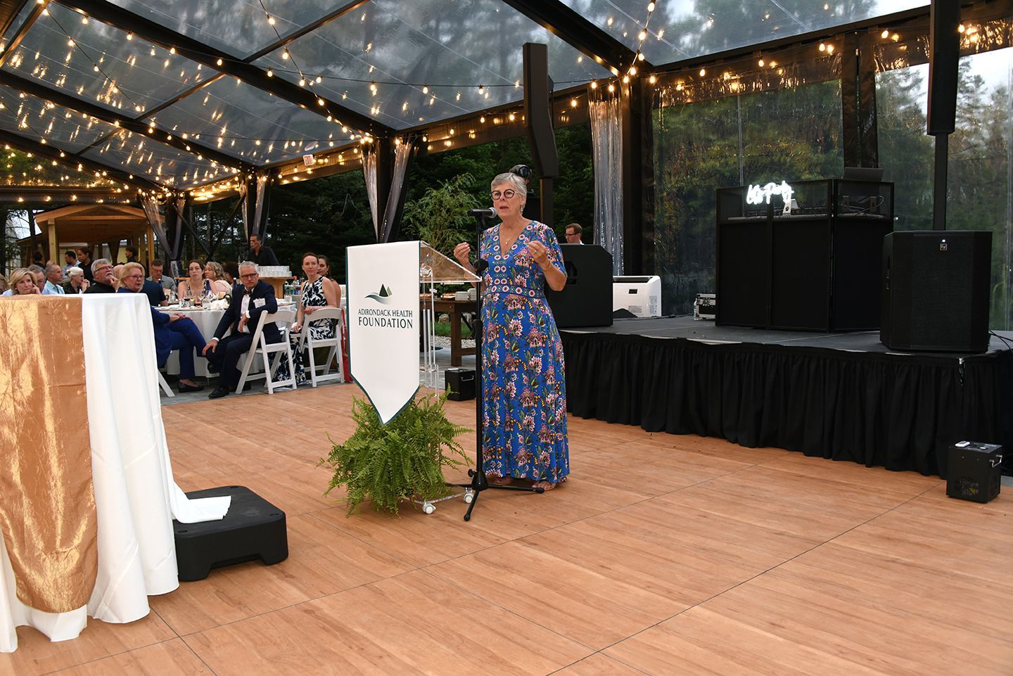 Woman speaking at an outdoor event under a clear tent, with a banner on the podium.