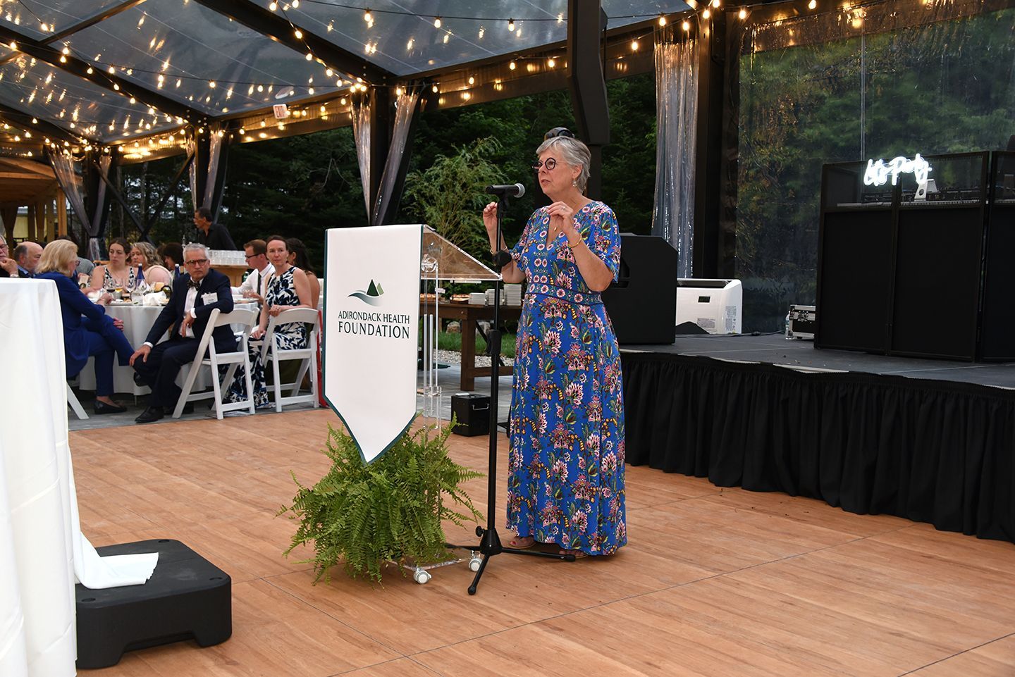 Woman speaking at an outdoor event, wearing a blue floral dress. Guests sit at tables in the background.
