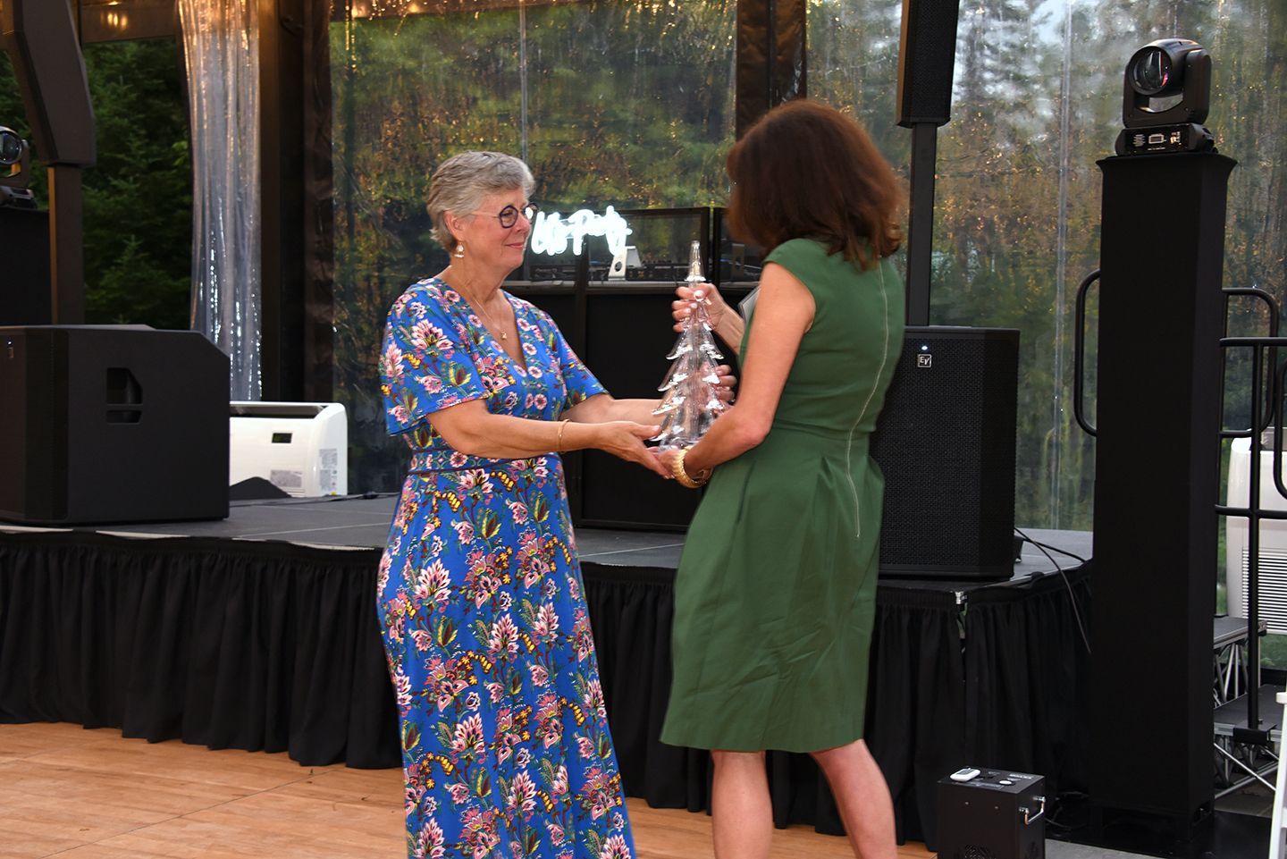 Woman in floral dress presents a silver trophy to a woman in a green dress on a stage. They are smiling.